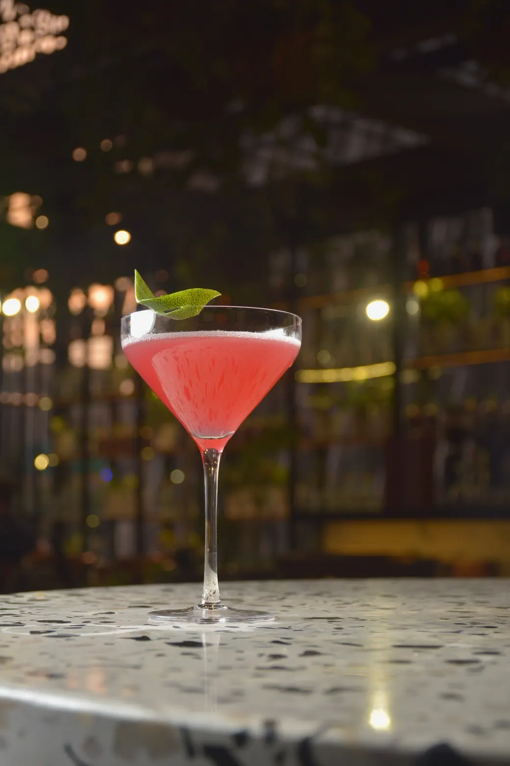 Coctel rosa en copa coupé con una hoja de limón, sobre una mesa de terrazo en Brick